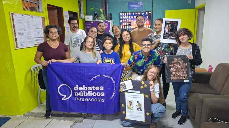 Grupo de jovens e adultos em uma sala colorida, posando com uma bandeira dos Debates Públicos nas Escolas e cartazes sobre direitos humanos.