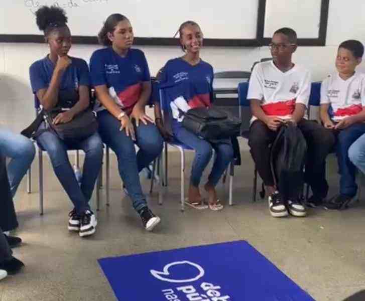 Grupo de cinco estudantes sentados em cadeiras dentro de uma sala, formando um círculo. Eles usam uniformes escolares nas cores azul, branco e vermelho e estão participando de um debate. No chão, ao centro, há a bandeira dos Debates Públicos.