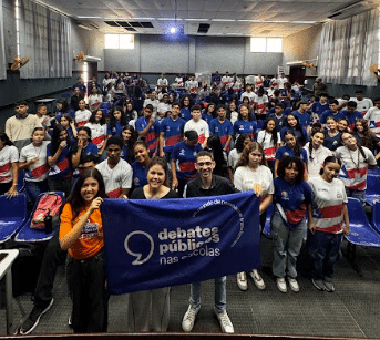 Um grande grupo de jovens reunidos em um auditório, posando para uma foto durante um encontro dos Debates Públicos nas Escolas. Eles aparecem sorrindo, enquanto algumas pessoas na frente seguram a bandeira dos Debates Públicos.