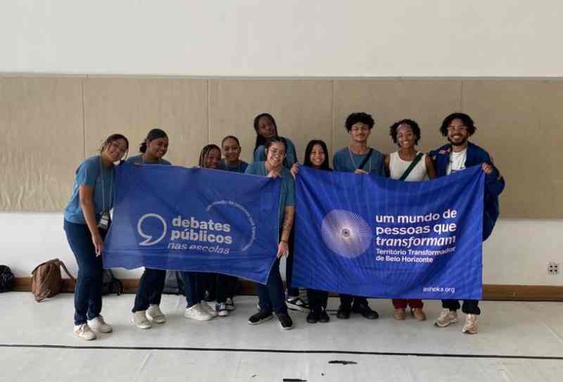 Foto de grupo tirada no Centro de Referência da Juventude, em Belo Horizonte/MG. No centro da imagem, pessoas seguram duas bandeiras azuis: uma com o logo dos Debates Públicos nas Escolas e outra do Território Transformador de Belo Horizonte
