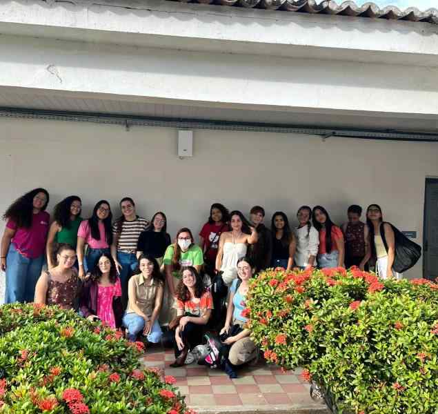 Grupo de mulheres reunidas, elas estão posicionadas lado a lado, sorrindo para a foto. Em frente a algumas delas, nos cantos inferior direito e inferior esquerdo da imagem, há duas moitas com flores alaranjadas.