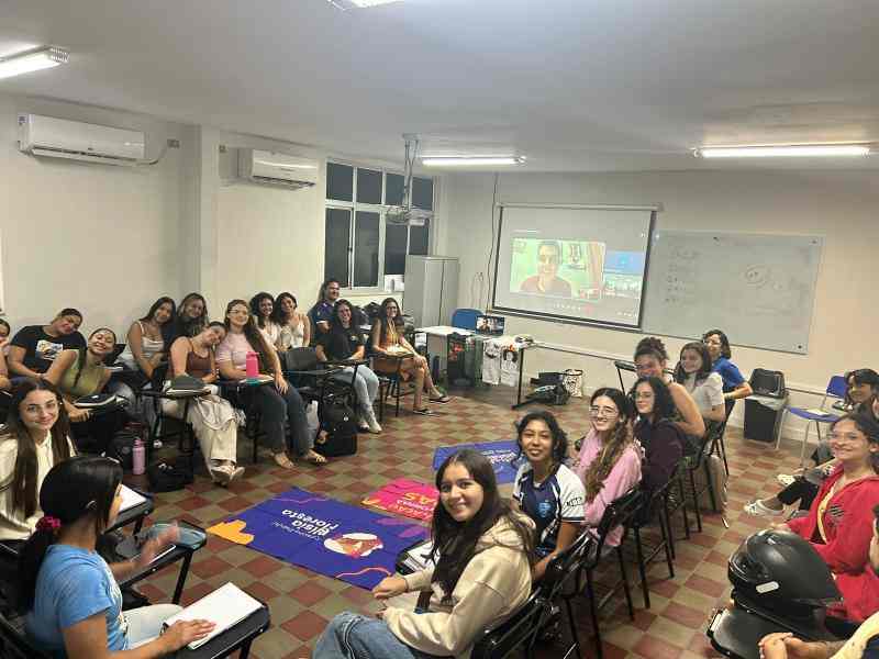 Grupo de estudantes reunidas em uma sala de aula, sentadas em semicírculo e sorrindo para a foto. No centro da sala há diferentes bandeiras dos Debates Públicos e do Cursinho Nísia Floresta, e ao fundo uma tela de projeção mostra uma pessoa participando por videoconferência.