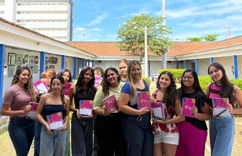 Grupo de onze jovens sorrindo em pátio de universidade, segurando cadernos rosa e roxo, com prédios brancos e colunas azuis ao fundo.